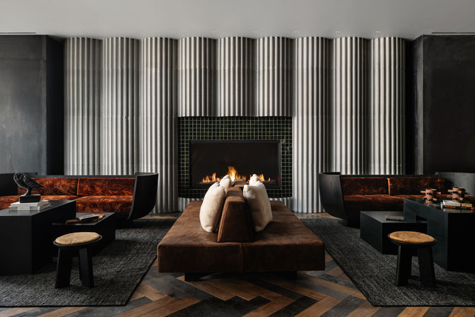 Contemporary luxury lounge with a central brown leather sofa and white pillows facing a glowing black‑tiled fireplace, flanked by rust velvet sofas, ribbed concrete wall panels and chevron wood flooring.