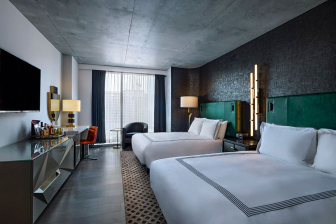 Stylish urban hotel room with two queen beds, green upholstered headboards against a dark textured wall, floor-to-ceiling window with sheer curtains and city view, mirrored desk with minibar, wall-mounted TV, bedside lamps and a seating area.