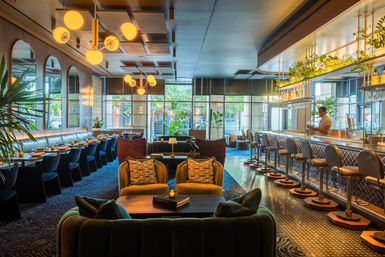 Chic urban hotel bar and lounge with velvet sofas and armchairs, brass globe pendant lights, long banquette dining, row of barstools at a tiled bar, hanging plants and large windows showing a sunlit city street.