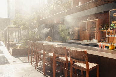 Sunlit urban rooftop patio bar with woven wooden bar stools along a concrete counter, potted plants and pergola seating, hazy sunbeams and colorful cocktails on the bar.