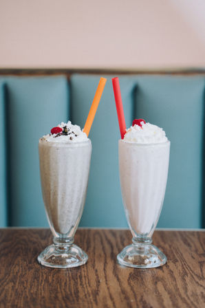 Two retro diner milkshakes in tall glass goblets topped with whipped cream and cherries, orange and red straws on a wooden table with a teal booth backdrop