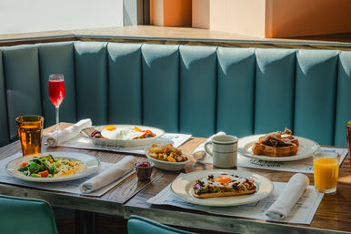 Sunlit brunch spread in a teal booth: plates with sunny-side-up eggs on toast and with roasted veggies, waffles with syrup, mixed salad, a bowl of golden fries, coffee mug, orange juice, and a red sparkling drink on a wooden table.