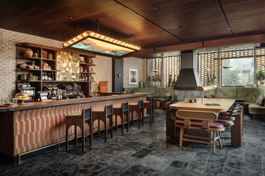 Stylish mid-century modern cafe interior with a long wooden bar and row of stools, glowing marquee light fixture, communal wooden table, fireplace, brick wall shelving and leafy potted plants.