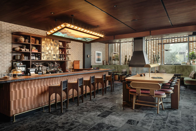 Stylish mid-century modern cafe interior with a long wooden bar and row of stools, glowing marquee light fixture, communal wooden table, fireplace, brick wall shelving and leafy potted plants.