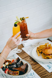 Bright cafe brunch: two hands toasting a blackberry-topped mimosa and a celery-and-olive Bloody Mary, with French toast and berries on one plate and a cheeseburger with fries on the other.