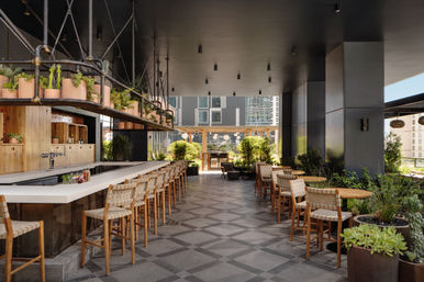 Modern rooftop bar and urban patio with a long white bar and woven wooden stools, round wood tables, hanging planters and potted greenery, open-air seating overlooking contemporary high-rise city buildings.