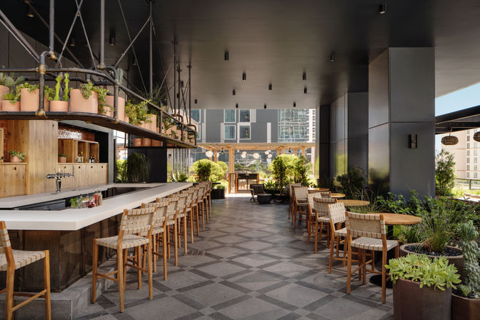 Modern rooftop bar and urban patio with a long white bar and woven wooden stools, round wood tables, hanging planters and potted greenery, open-air seating overlooking contemporary high-rise city buildings.