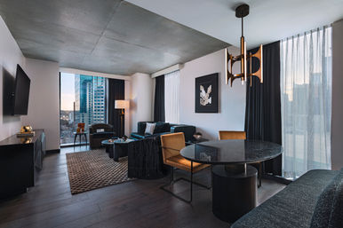 Stylish modern high-rise living-dining room with floor-to-ceiling windows overlooking a downtown skyline, concrete ceiling, teal sofa, round marble table, and mustard chairs.