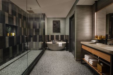 Modern spa-style bathroom with freestanding stone bathtub against a textured gray wall, glass-enclosed walk-in shower with dark vertical tiles on the left, long vanity with illuminated mirror and open wood shelving on the right, and speckled terrazzo flooring.