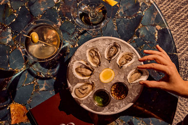 Top-down shot of a chilled oyster platter on crushed ice with lemon wedge and mignonette sauces, a hand reaching for an oyster and a martini with an olive on a blue marble table.