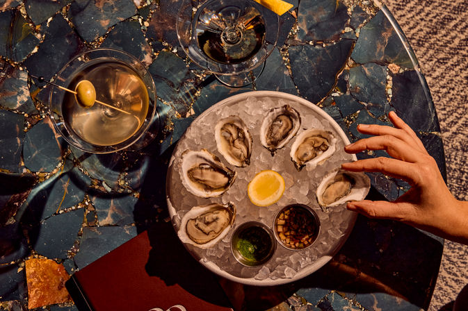Top-down shot of a chilled oyster platter on crushed ice with lemon wedge and mignonette sauces, a hand reaching for an oyster and a martini with an olive on a blue marble table.