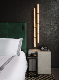 Modern bedroom corner with emerald green upholstered headboard, crisp white bedding with black stripes, marble nightstand and black tray holding a coffee cup and books, retro-style radio, and warm vertical LED wall sconces against a dark textured wall.