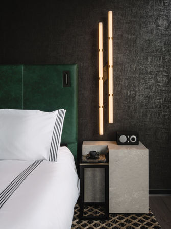 Modern bedroom corner with emerald green upholstered headboard, crisp white bedding with black stripes, marble nightstand and black tray holding a coffee cup and books, retro-style radio, and warm vertical LED wall sconces against a dark textured wall.