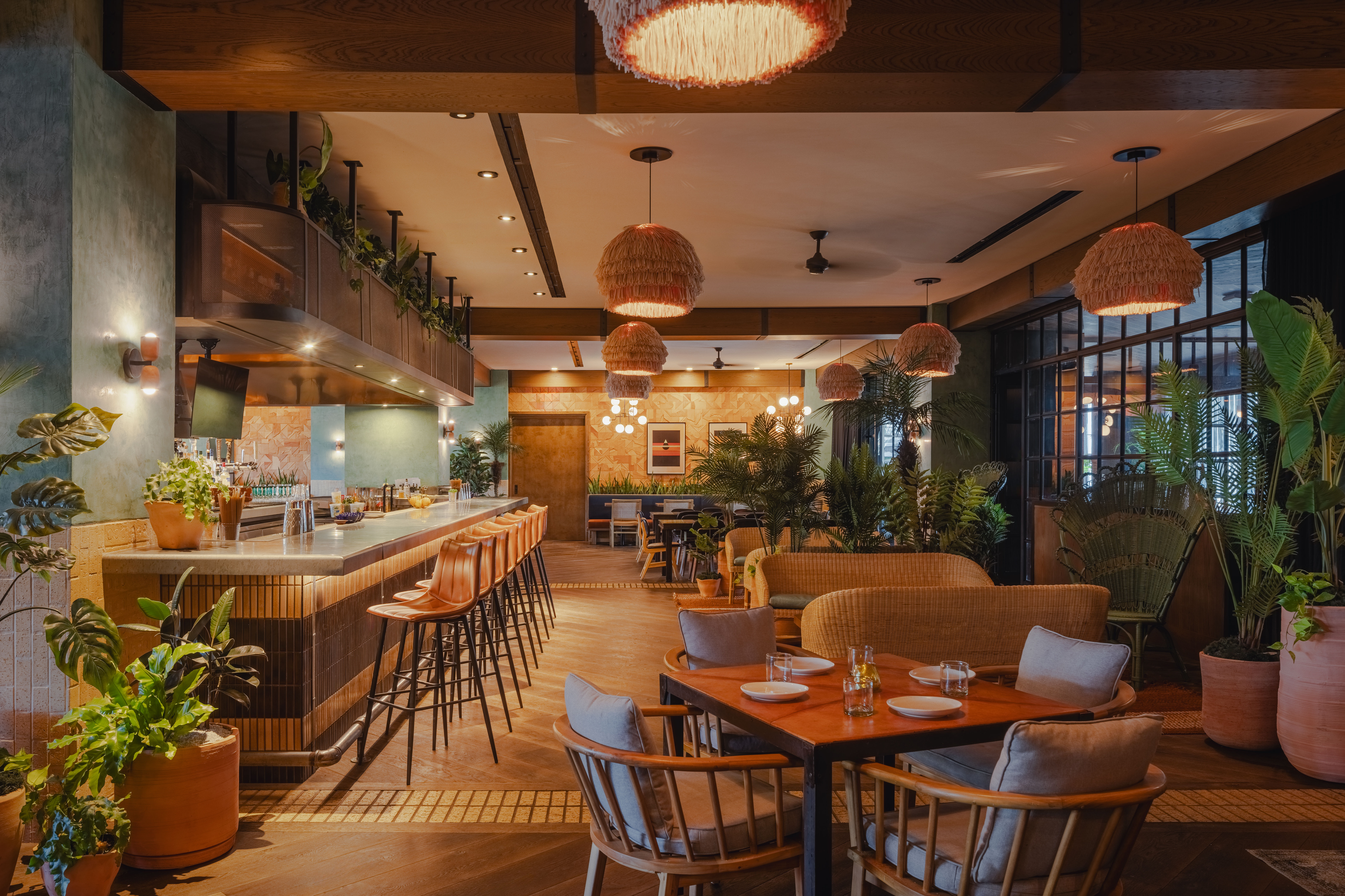 Cozy modern boho restaurant interior with rattan pendant lights, a long bar with leather stools, wooden dining tables, wicker lounge seating, abundant potted plants, and warm ambient lighting.