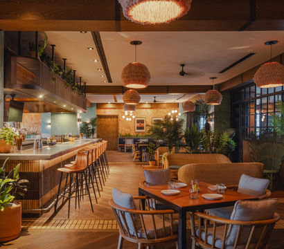 Cozy modern boho restaurant interior with rattan pendant lights, a long bar with leather stools, wooden dining tables, wicker lounge seating, abundant potted plants, and warm ambient lighting.