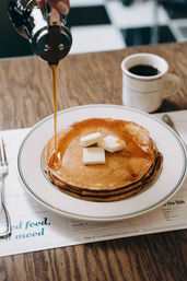 Stack of fluffy pancakes topped with pats of butter while maple syrup is poured over them, served with a mug of black coffee on a wooden diner table.
