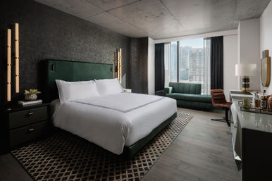 Stylish modern hotel room with king bed and green upholstered headboard, dark textured accent wall, vertical pendant lights, patterned rug, green sofa by floor‑to‑ceiling window with city skyline view, desk and minibar.
