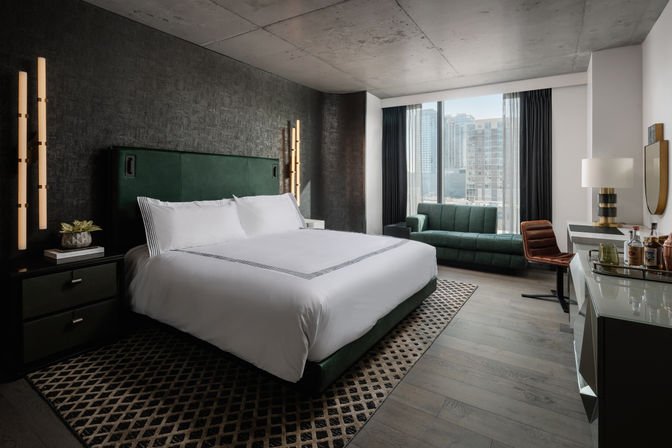 Stylish modern hotel room with king bed and green upholstered headboard, dark textured accent wall, vertical pendant lights, patterned rug, green sofa by floor‑to‑ceiling window with city skyline view, desk and minibar.
