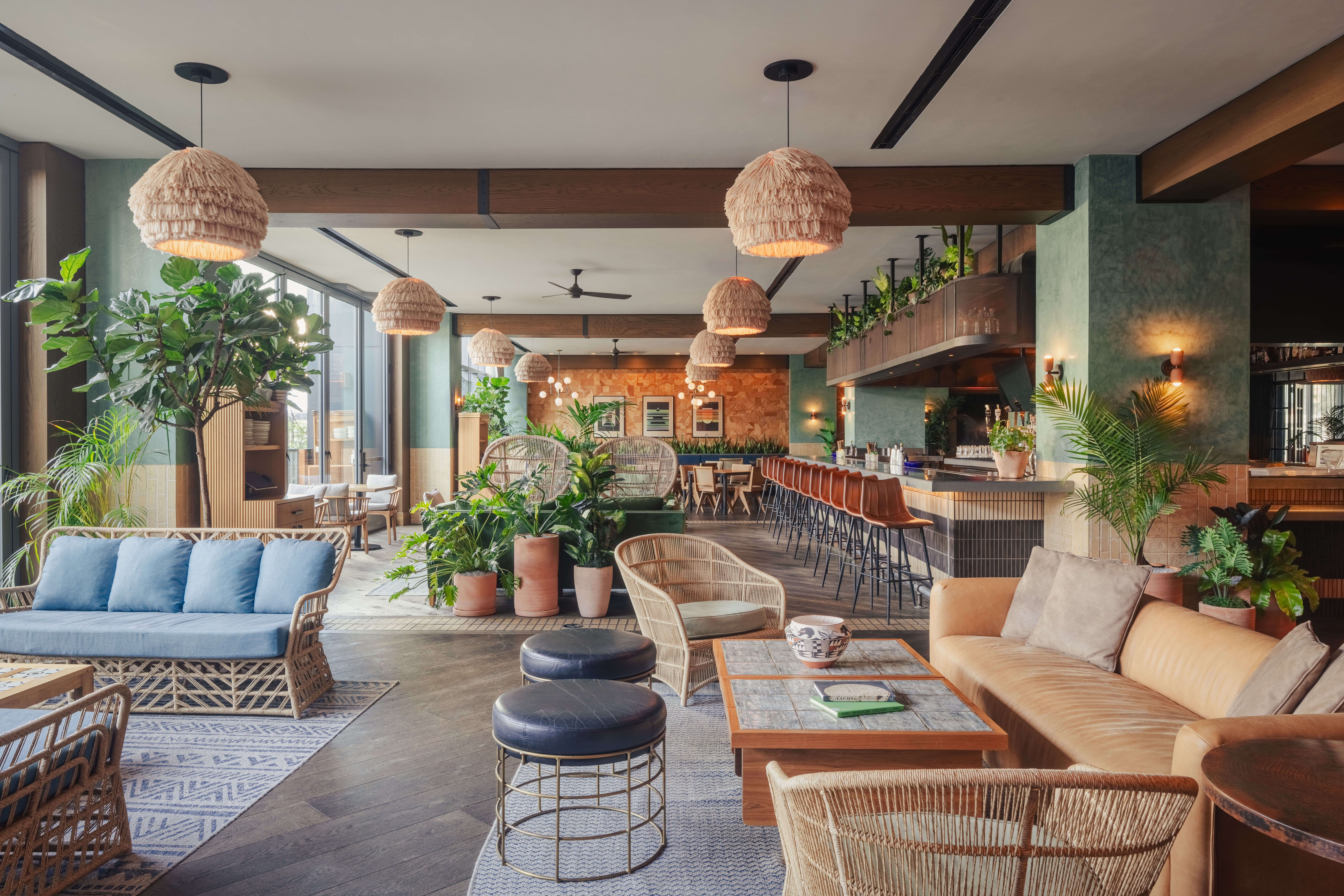 Bright tropical-inspired hotel lounge and bar with rattan sofas, blue cushions, woven pendant lights, leather bar stools and abundant potted plants