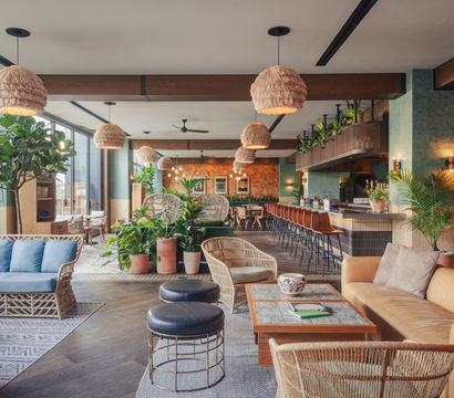 Bright tropical-inspired hotel lounge and bar with rattan sofas, blue cushions, woven pendant lights, leather bar stools and abundant potted plants