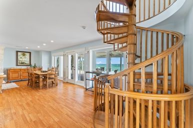 Beachfront interior with a wooden spiral staircase, warm hardwood floors, rustic dining table and glass doors framing turquoise ocean views.