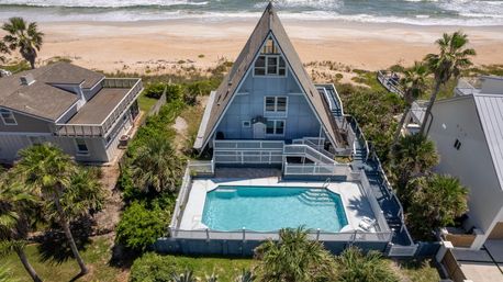 Aerial view of a beachfront A-frame house with a fenced turquoise pool, palm trees and sandy ocean shoreline