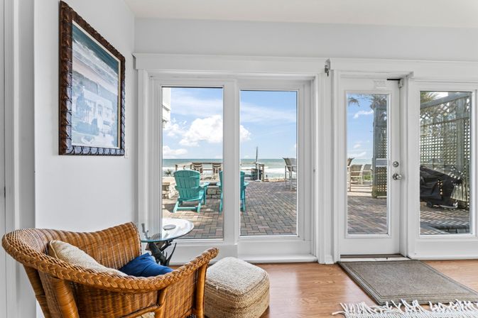 Sunlit coastal living room with wicker chair and ottoman by glass French doors opening to a brick beachfront patio with turquoise Adirondack chairs, outdoor seating, and ocean view under a blue sky.