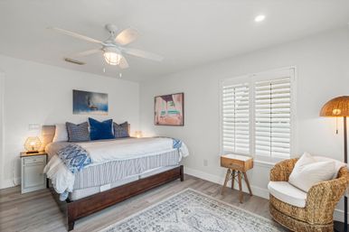 Bright coastal-style guest bedroom with dark wood bed dressed in white and blue linens, navy accent pillows, ceiling fan, plantation shutters, woven rattan armchair, bedside tables and warm ambient lamps