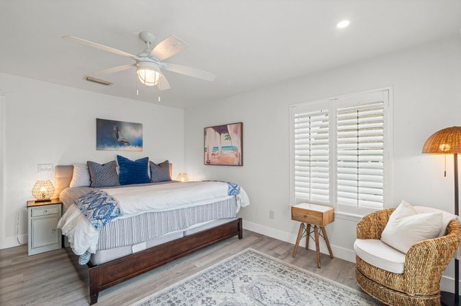 Bright coastal-style guest bedroom with dark wood bed dressed in white and blue linens, navy accent pillows, ceiling fan, plantation shutters, woven rattan armchair, bedside tables and warm ambient lamps
