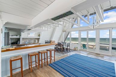 Sunlit beachfront open-plan living room and kitchen with white vaulted beams, wooden bar with four stools, dining table, blue striped rug, chandelier, and floor-to-ceiling windows opening to a sandy beach and ocean view.