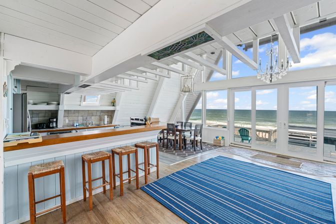 Sunlit beachfront open-plan living room and kitchen with white vaulted beams, wooden bar with four stools, dining table, blue striped rug, chandelier, and floor-to-ceiling windows opening to a sandy beach and ocean view.