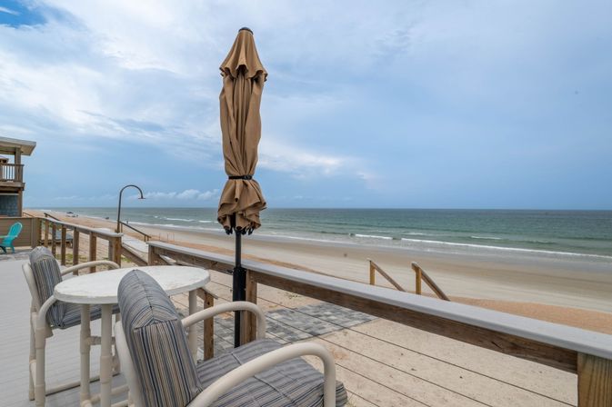 Relaxing oceanfront deck with striped cushioned chairs, round table and closed umbrella overlooking a wide sandy beach and gentle waves under a cloudy blue sky.