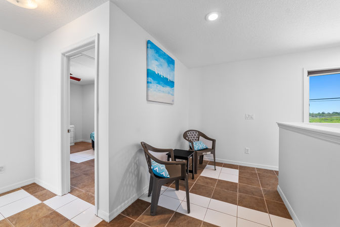 Cozy upstairs landing seating nook with two brown chairs and blue chevron pillows, small black side table, coastal blue canvas on a white wall, and brown-and-white tiled floor with a window view of green landscape.