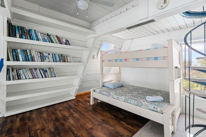 Bright coastal loft bedroom with white shiplap walls, built-in bookshelves filled with books, dark hardwood floor, white twin-over-twin bunk bed with patterned bedding, and a spiral staircase opening to a balcony with palm trees and blue sky.