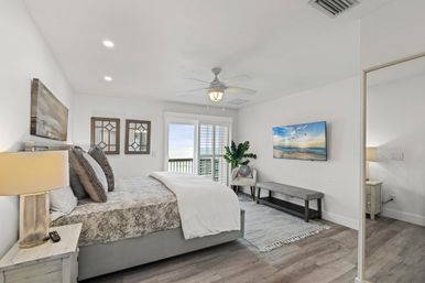 Bright coastal bedroom with ocean-view balcony, neutral palette, large bed with textured bedding, ceiling fan, wall-mounted TV, bench, indoor plant and sliding glass doors flooding the room with natural light.