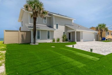 Two-story coastal beach house with light gray siding, palm trees and bright green lawn, paved driveway and two-car garage, blue ocean and clear sky in the background.