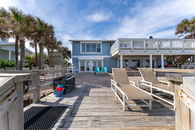 Coastal blue beach house with raised white deck, sun loungers on a weathered wooden boardwalk, palm trees and a patio with turquoise chairs under a bright sky.