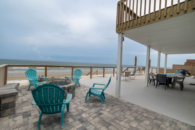 Oceanfront beach deck with turquoise Adirondack chairs around a stone fire pit, paved patio and covered dining area overlooking sandy shore and calm ocean under a cloudy sky.