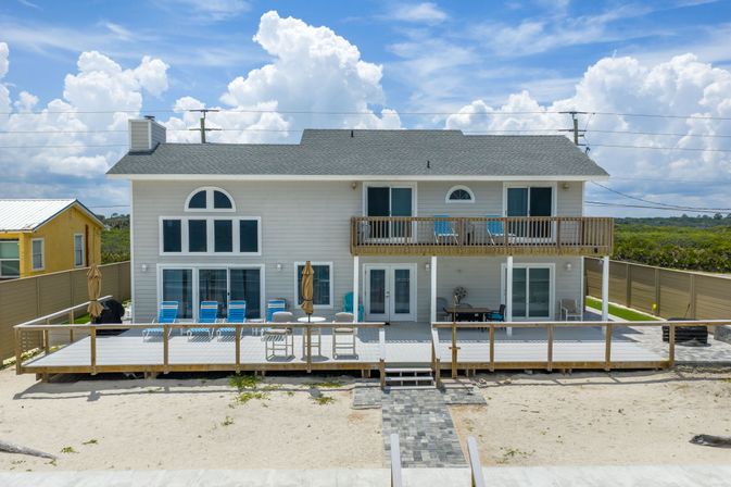 Two-story beachfront house with wide wooden deck and upper balcony, blue lounge chairs on sand under puffy white clouds