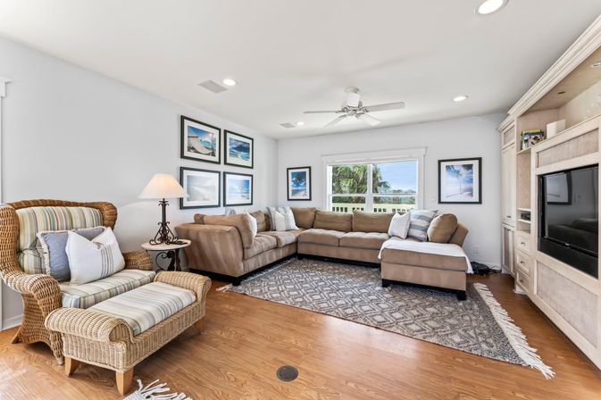 Sunny coastal living room with a large L-shaped beige sectional, wicker armchair and ottoman with striped cushions, patterned area rug on hardwood floors, built-in entertainment center, framed beach photos, and a window view of palm trees and blue sky.