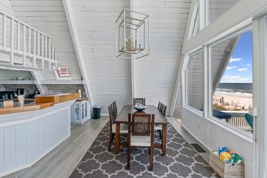 Bright coastal A-frame dining area with wooden table and chairs, geometric pendant light, open kitchen bar, patterned rug and large windows framing a sandy beachfront and ocean view.