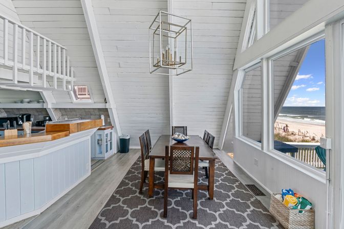Bright coastal A-frame dining area with wooden table and chairs, geometric pendant light, open kitchen bar, patterned rug and large windows framing a sandy beachfront and ocean view.