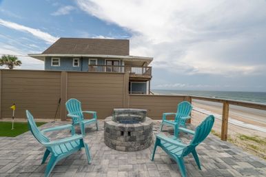 Beachfront paver patio with round stone fire pit surrounded by five turquoise Adirondack chairs, fenced deck and sandy ocean shoreline under a cloudy sky.