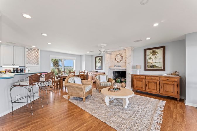Bright, open-concept coastal living room and kitchen with wicker armchairs, round coffee table on a textured rug, stone fireplace, wooden sideboard, bar stools, dining area and a window view of a palm tree and water.