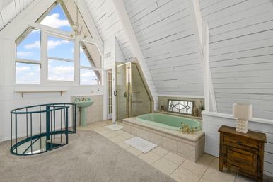 Sunny A-frame loft bathroom with large triangular windows, mint-green soaking tub with brass fixtures, glass shower, pedestal sink, spiral metal staircase, white shiplap walls