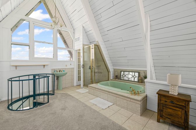 Sunny A-frame loft bathroom with large triangular windows, mint-green soaking tub with brass fixtures, glass shower, pedestal sink, spiral metal staircase, white shiplap walls