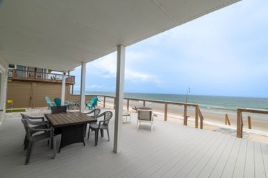 Covered beachfront deck with dining table, mixed plastic and wicker chairs, turquoise Adirondack seats and a lounge chair overlooking a sandy beach and calm ocean under a cloudy sky