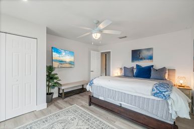 Bright, beachy master bedroom with king bed in blue-and-white linens, navy throw pillows, wooden platform frame, ceiling fan, wall-mounted TV showing a seaside scene, bench and potted plant by the closet, woven bedside lamps and light wood flooring.