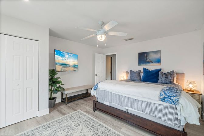 Bright, beachy master bedroom with king bed in blue-and-white linens, navy throw pillows, wooden platform frame, ceiling fan, wall-mounted TV showing a seaside scene, bench and potted plant by the closet, woven bedside lamps and light wood flooring.