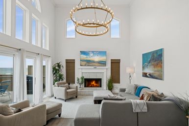 Sunlit coastal living room with vaulted ceilings and double-ring chandelier, fireplace with TV, gray sectional and armchairs, large windows and sliding doors opening to an ocean-view balcony.